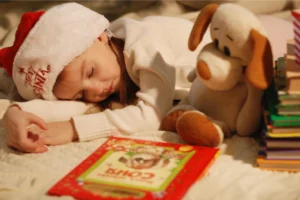 un bambino che dome con un libro a finaco e un cappello di babbo natale